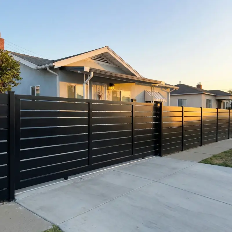 A Modern Black Metal Fence With Horizontal Slats And A Sliding Driveway Gate, Positioned In Front Of A SingleStory Stucco House Under A Clear Morning Sky