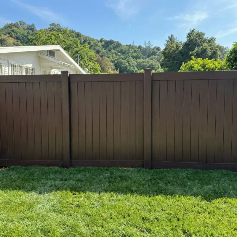 Dark Walnut Brown Vinyl Privacy Fence Installed In A Residential Backyard With Lush Green Grass And A Hillside Background