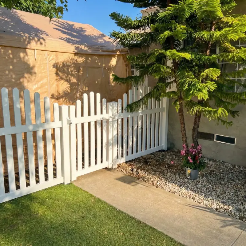 Green Artificial Grass Lawn In A Sunny Backyard, Bordered By A White Fence And Mature Palm Trees