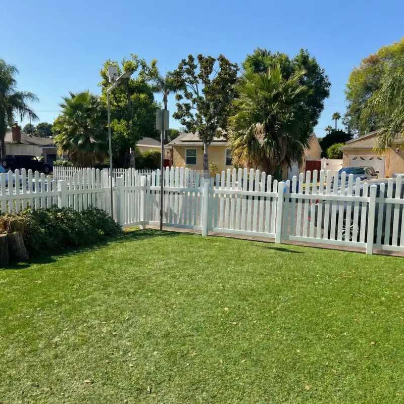 Sunny Residential Backyard With Green Artificial Grass, A White Vinyl Picket Fence, And A Yellow House With Palm Trees In The Background