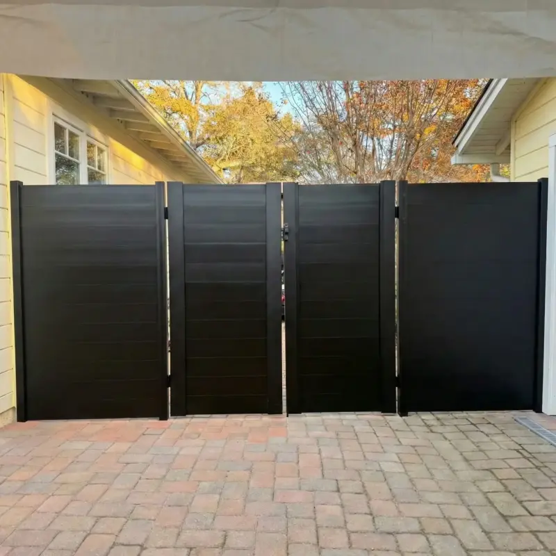 Tall Black Horizontal Plank Driveway Gate And Fence Panels Screening A Backyard, Situated On Interlocking Brick Pavers Next To A House With Yellow Siding