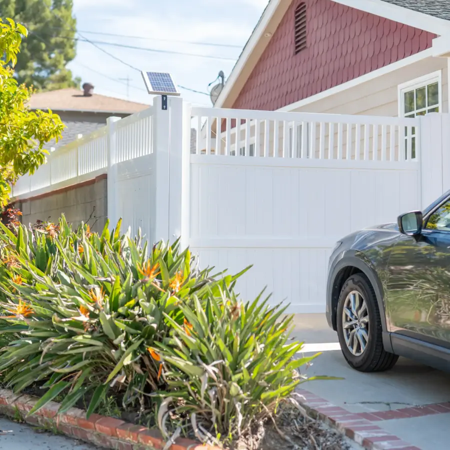 A Closed White Vinyl Gate Across A Driveway Next To A House, With A Dark Suv Parked Nearby And Lush Plants In The Foreground.