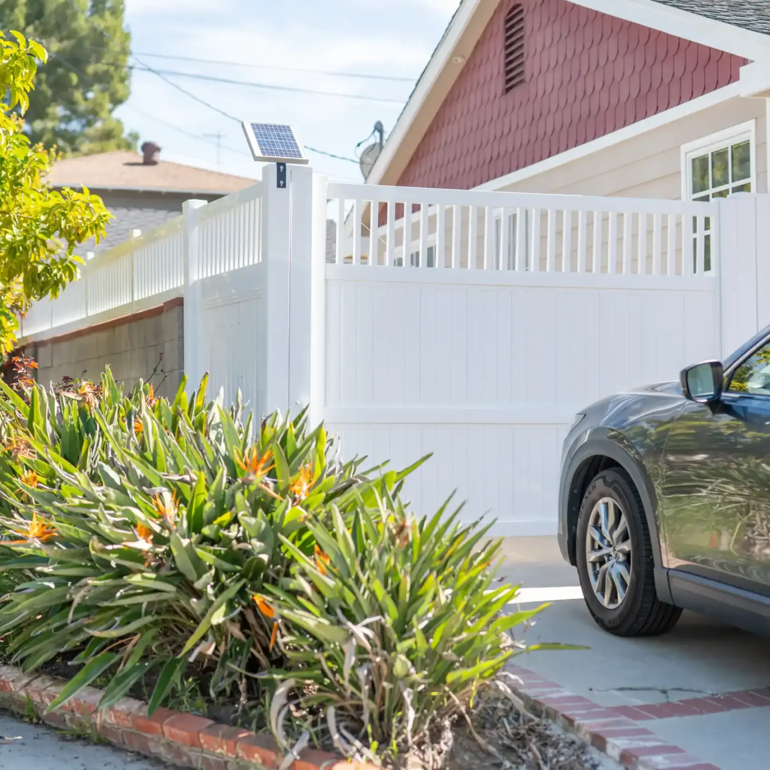 A closed white vinyl gate across a driveway next to a house, with a dark SUV parked nearby and lush plants in the foreground.
