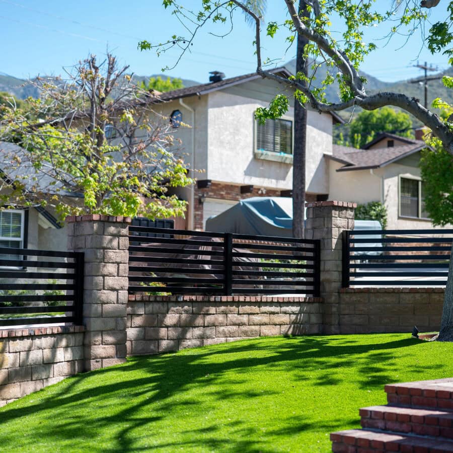Modern Black Horizontal Slat Fencing Installed On Top Of A Tan Stone Retaining Wall With Brick Trim, Set In A Sunny Suburban Front Yard With Bright Green Grass And A Large Tree.