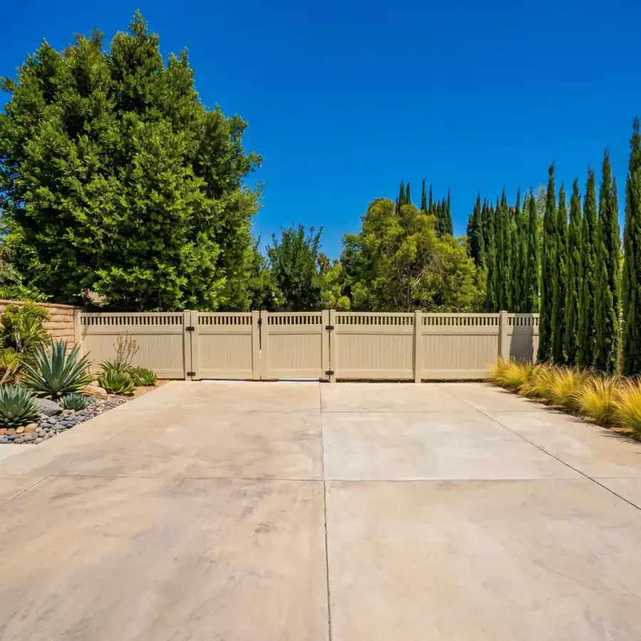 A Wide Concrete Driveway Leading To A Beige Privacy Fence Surrounded By Lush Green Trees And Landscaped Desert Plants Under A Clear Blue Sky.