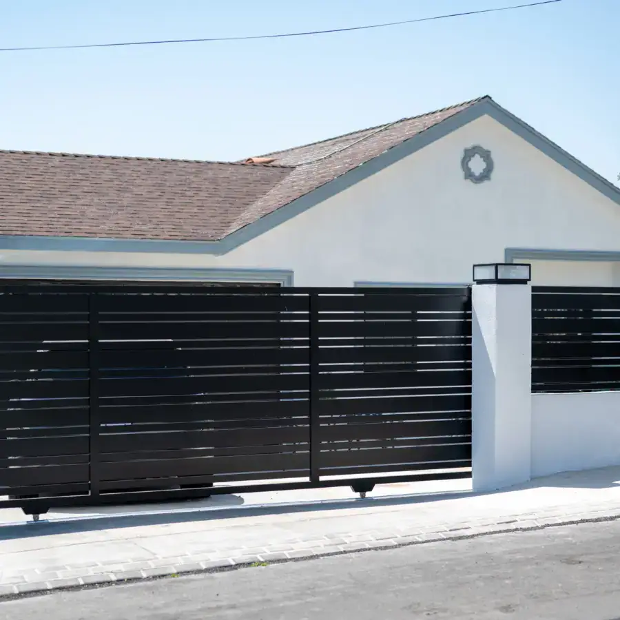 A Modern Black Horizontal Slat Sliding Driveway Gate, Partially Open, In Front Of A Beige Stucco Home With An Open Garage. The Gate Is Supported By StoneCapped Pillars Under A Bright Blue Sky, With A Paved Street In The Foreground.