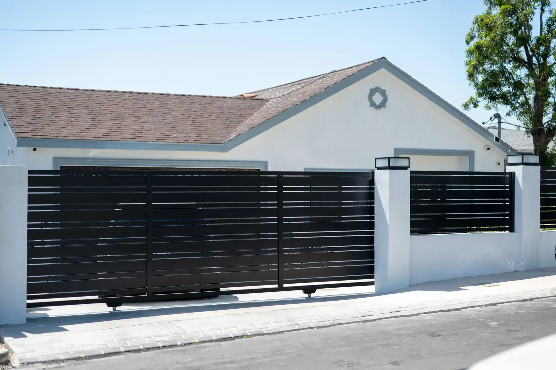 A Modern Black Horizontal Slat Sliding Driveway Gate, Partially Open, In Front Of A Beige Stucco Home With An Open Garage. The Gate Is Supported By StoneCapped Pillars Under A Bright Blue Sky, With A Paved Street In The Foreground.