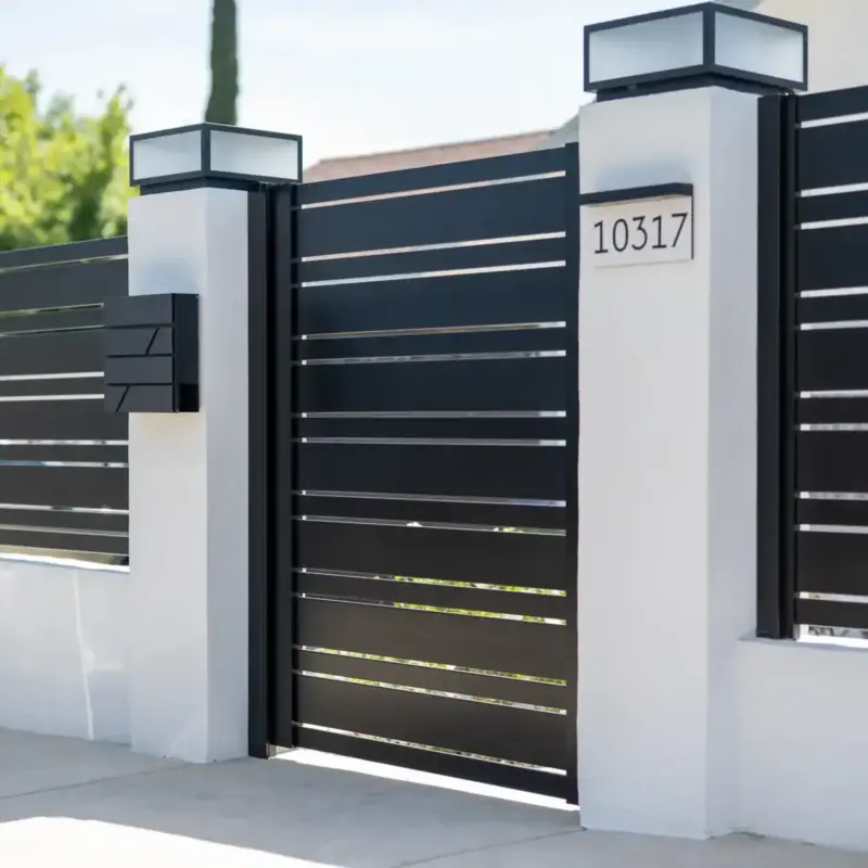 A Modern Residential Front Gate And Fence Made Of Black Horizontal Slats Between Two White Stucco Pillars, Each Topped With A Square Light. The Right Pillar Displays The House Number &Quot;10317&Quot;, And The Left Has A Black Slatted Mailbox. Concrete Sidewalk With Pavers Is In The Foreground Under Sunny Skies.