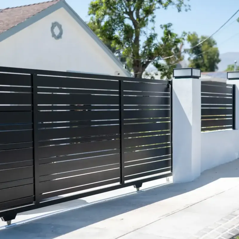 A Modern Black Horizontal Slat Sliding Driveway Gate, Partially Open, In Front Of A Beige Stucco Home With An Open Garage. The Gate Is Supported By StoneCapped Pillars Under A Bright Blue Sky, With A Paved Street In The Foreground.