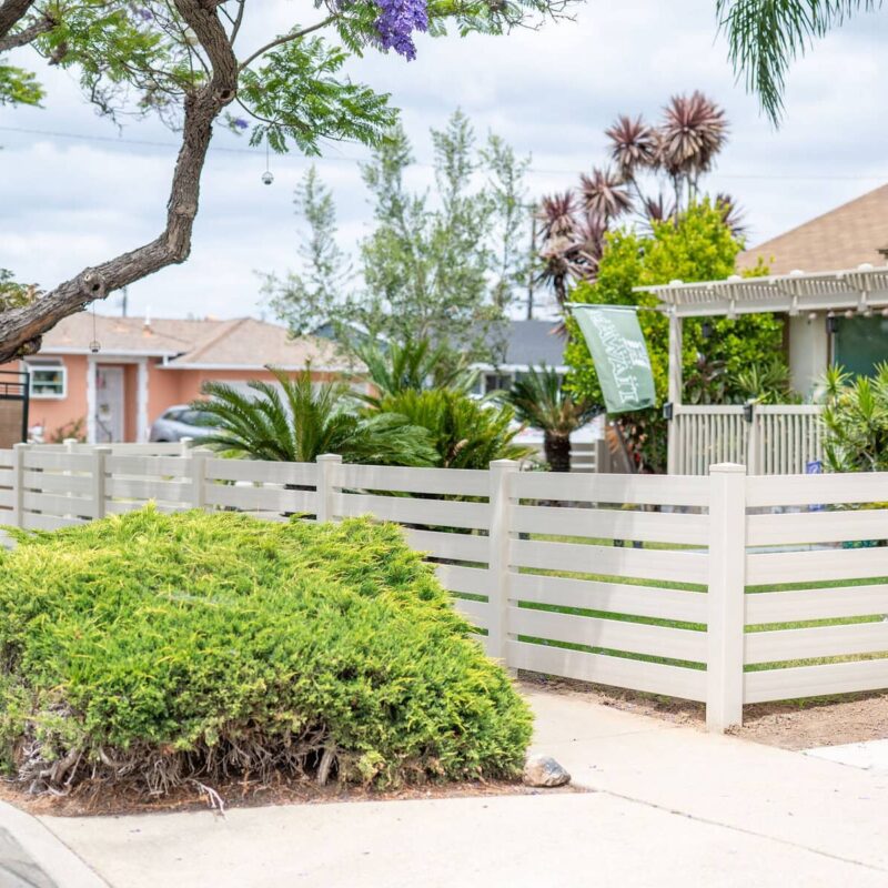 A Tan Vinyl Privacy Fence Featuring A Decorative Horizontal Slat Lattice Top, Installed Along A Concrete Sidewalk.