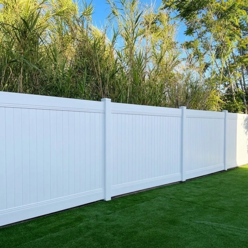 A Pristine White Vinyl Privacy Fence Installed In A Residential Backyard, Featuring Bright Green Artificial Turf And Backed By Lush Greenery Under A Clear Blue Sky.