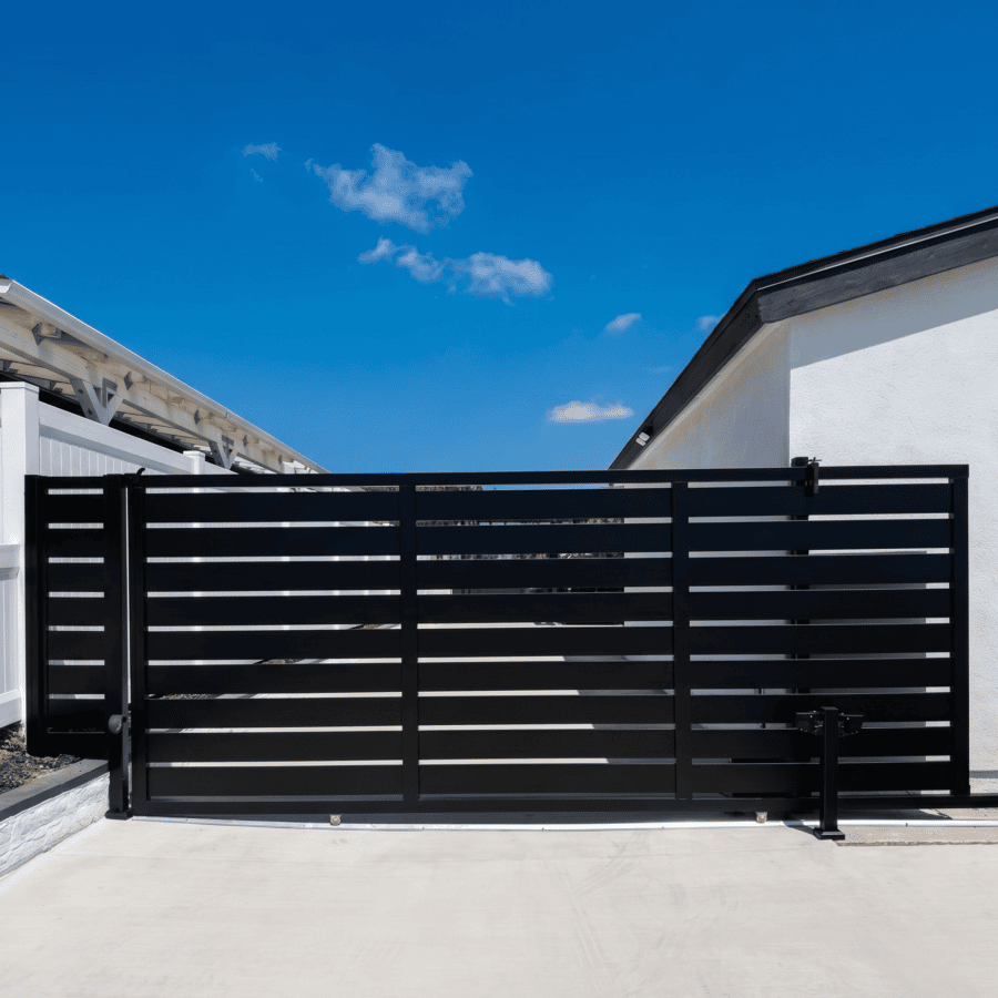 A Centered Photograph Of A Modern, Black Horizontal Slat Sliding Gate Across A Concrete Driveway, Set Between A White Vinyl Fence And A White Stucco Building, Under A Clear Blue Sky.