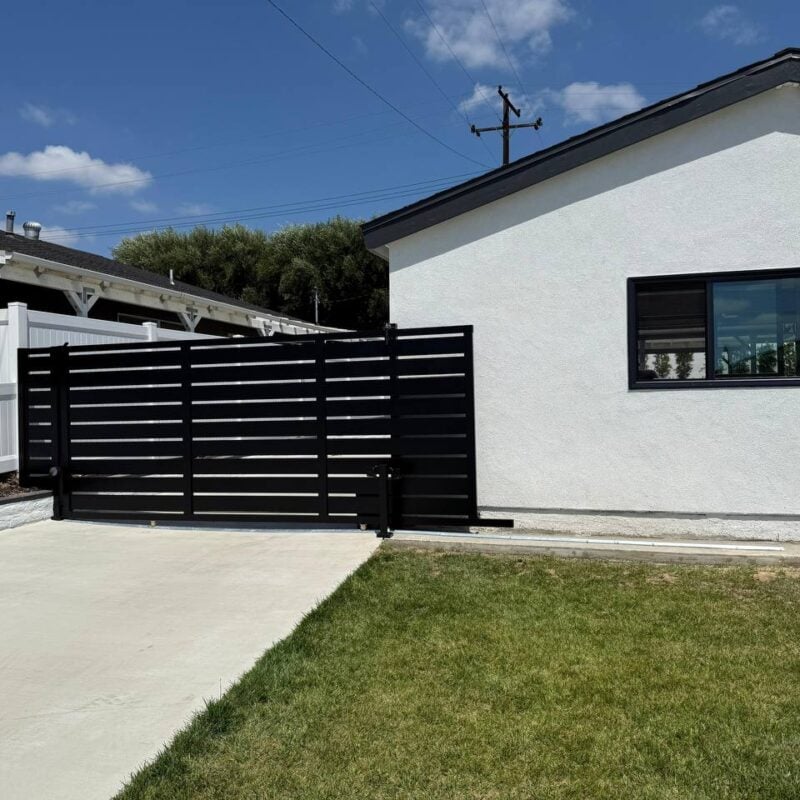 A Centered Photograph Of A Modern, Black Horizontal Slat Sliding Gate Across A Concrete Driveway, Set Between A White Vinyl Fence And A White Stucco Building, Under A Clear Blue Sky.