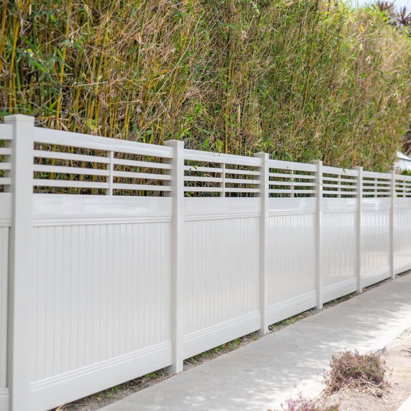 A Tan Vinyl Privacy Fence Featuring A Decorative Horizontal Slat Lattice Top, Installed Along A Concrete Sidewalk.