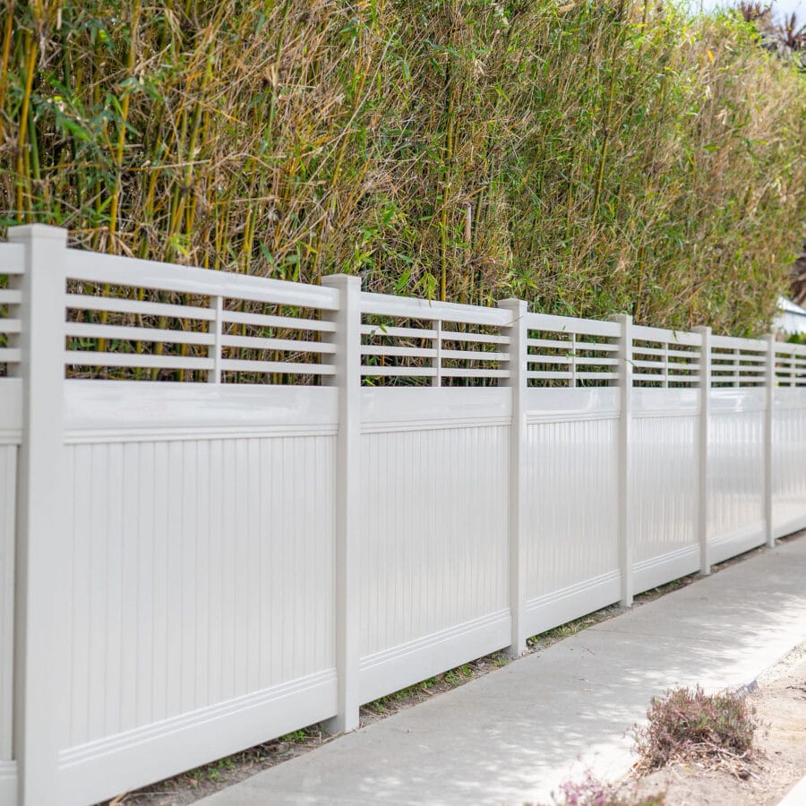 A Tan Vinyl Privacy Fence Featuring A Decorative Horizontal Slat Lattice Top, Installed Along A Concrete Sidewalk.