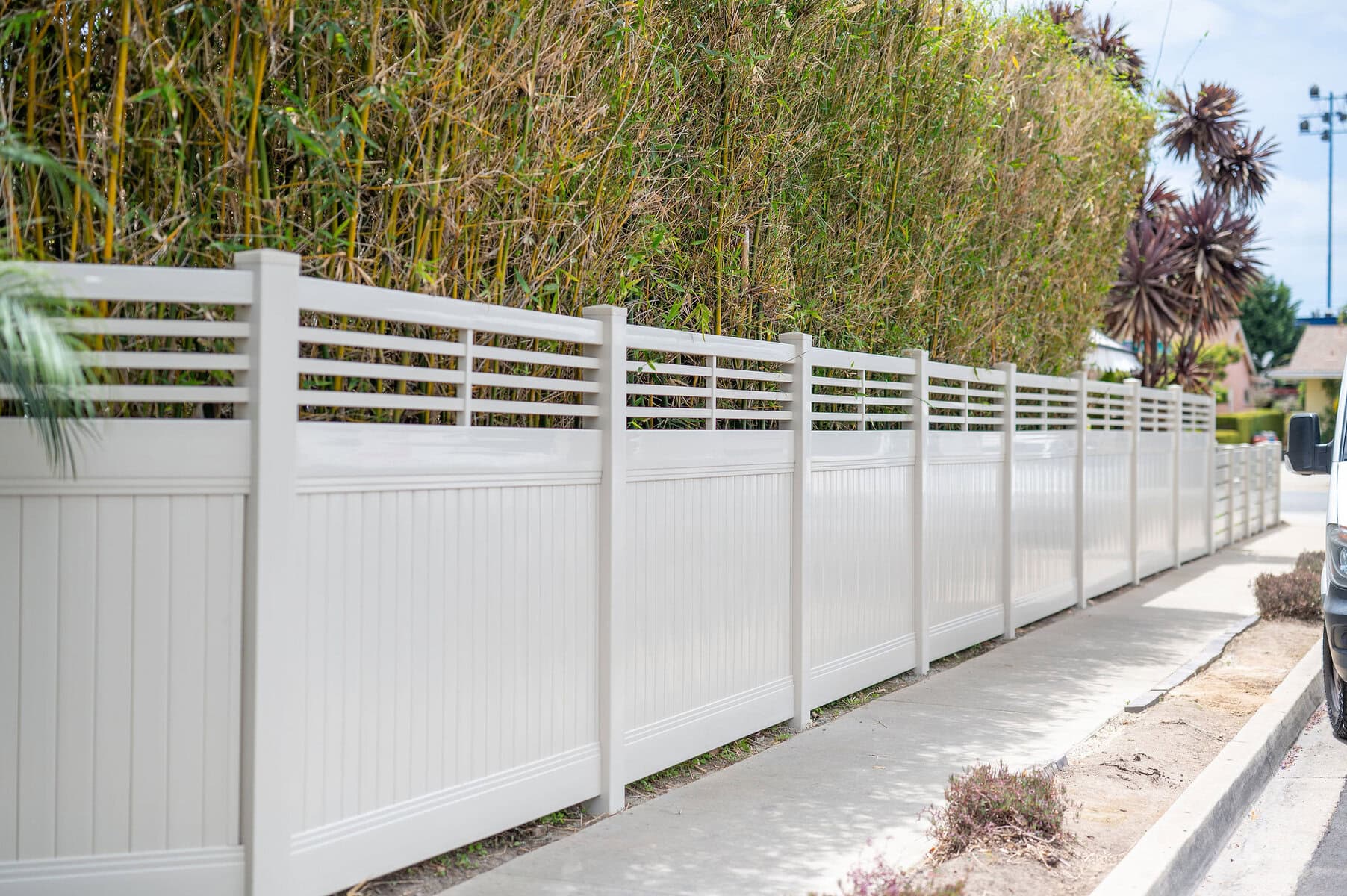 A Tan Vinyl Privacy Fence Featuring A Decorative Horizontal Slat Lattice Top, Installed Along A Concrete Sidewalk.